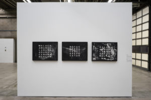 3 paintings hang on a white wall. The paintings are black with white letters on them, arranged in a "Magic Square".