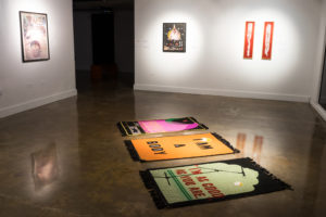 This photo shows the exhibition room containing rugs on the floor and 3 artworks hung on the wall.