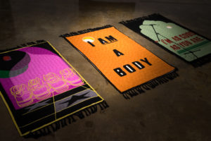 3 prayer rugs lay on the floor side by side. The first depicts Nike shoes reading "I am a Muslima", the second rug isays "I am a Body", and the third reads "I'm good as you are"