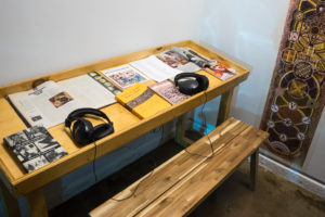 The photo depicts a bench sitting near an area that has 2 headphones, books, magazines, and other reading materials related to the artist Harry E. Smith