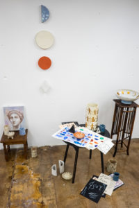 Installation composed of ceramics, paintings, and papers set on chairs.