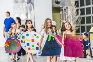 Children in vibrant costumes parade down a hallway, showcasing their colorful attire and joyful expressions.
